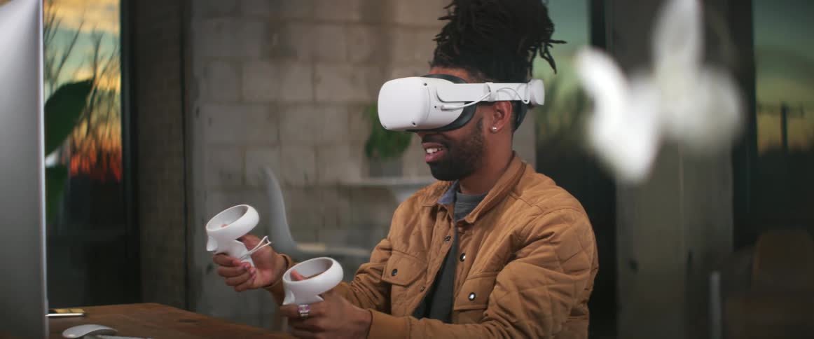 A black man sitting at a desk is wearing a virtual reality headset and holding the game controllers in his hands