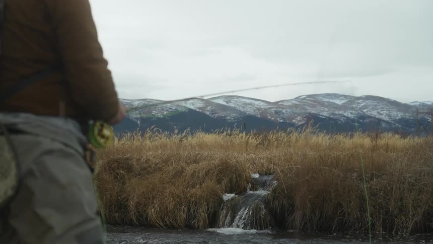 A man fishes in a river near a mountain range