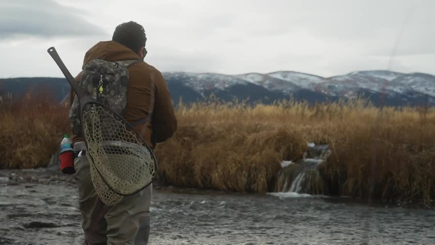 Man casts fishing line into river across from small waterfall flowing through marsh grass into water