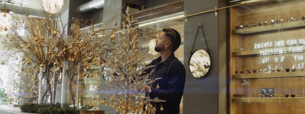 A man is standing inside a store hanging holiday ornaments on tree branches