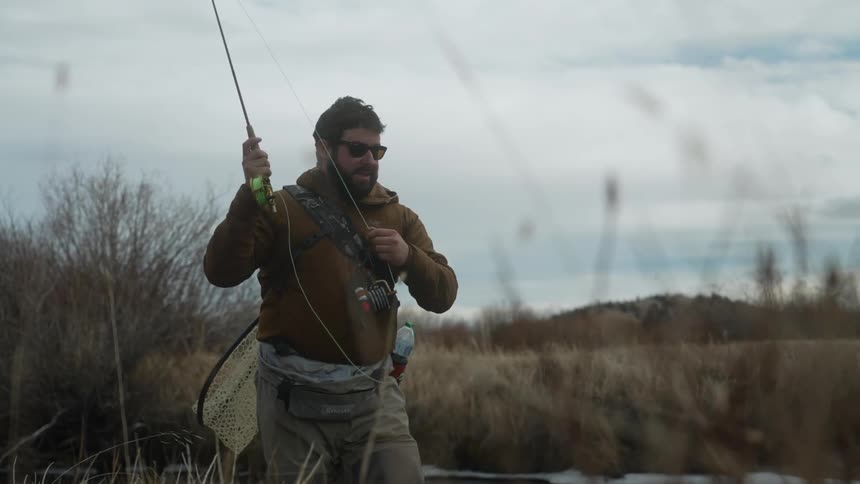 A fisherman is standing on a river bank near bare trees and casting his line