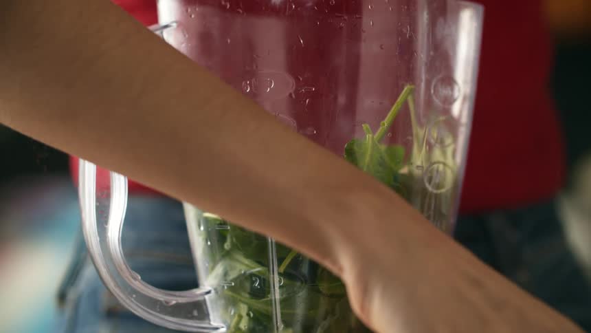 A woman adding sliced banana to spinach in a blender