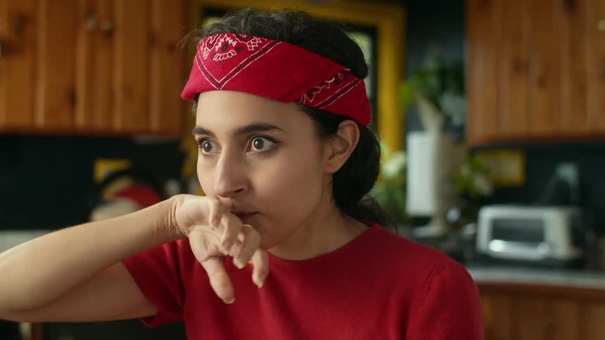 A teenage girl with long dark hair and a red bandana on her head is wide eyed as she drinks a glass of juice wipes her mouth and smiles