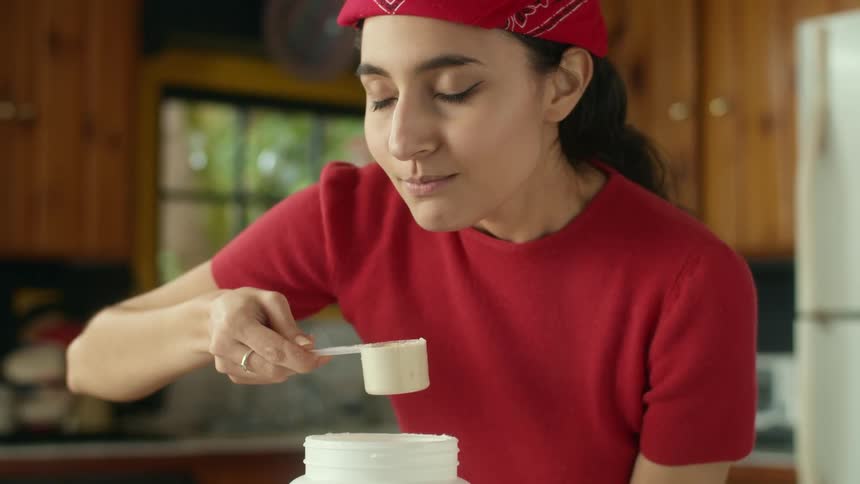 A teenage girl is in a kitchen putting a cup inside a large container and smelling what is inside