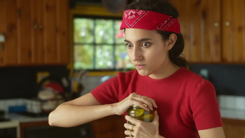 A teenage girl with long dark hair and a red bandana on her head is holding a pickle jar and making faces as she removes the lid