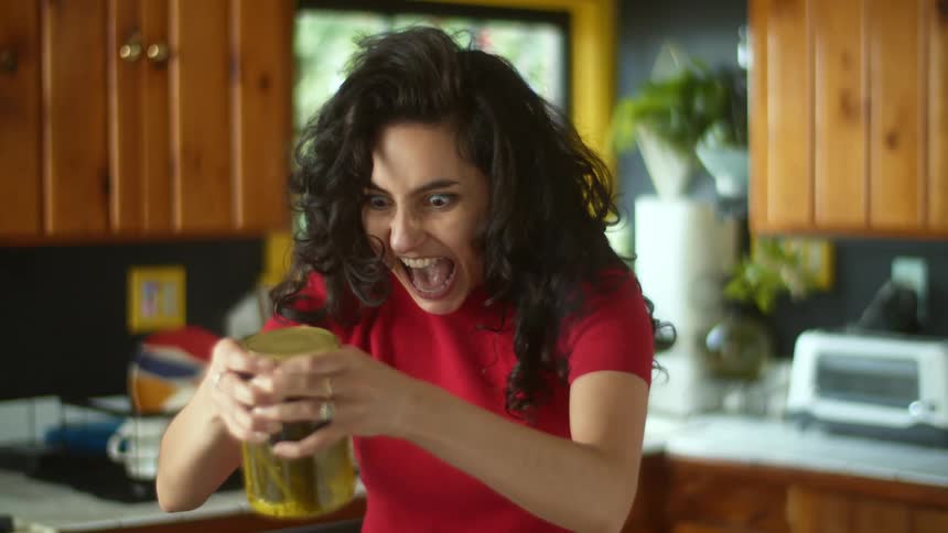 A teenage girl is standing in a kitchen holding a jar of pickles and screaming as she looks at it