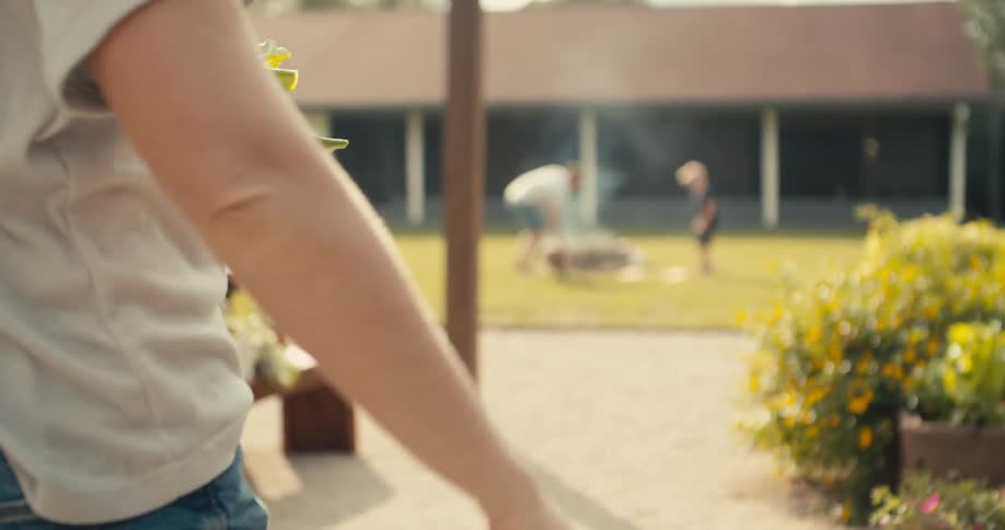 Woman carries potted plant past planters toward husband and son standing around fire pit in backyard