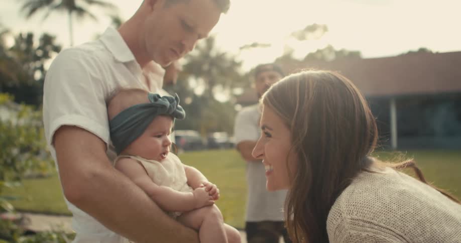 Father with polo shirt holds baby daughter and woman with wavy hair talks to baby in garden