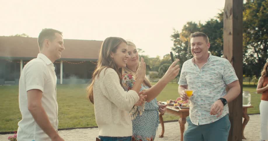 Friends stand outside at backyard picnic video calling on mobile phone