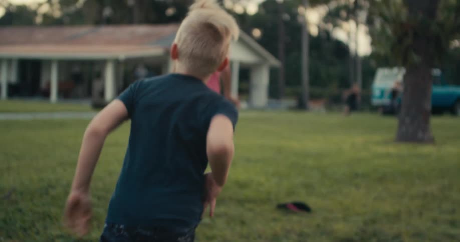 Blonde girl with pink t shirt and beige shorts runs barefoot with blonde boy wearing green t shirt and black shorts and sneakers chasing her on lawn in the garden