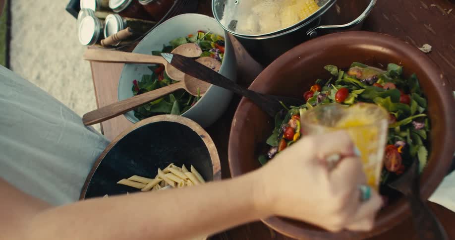 Person drizzles dressing over salad sitting on picnic table with other food