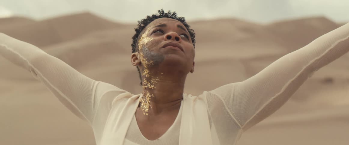 A woman with gold makeup on one side of her face outstretches her arms in the desert