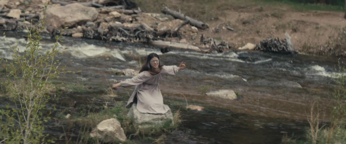 A woman is sitting on a rock in a stream and doing arm movements as the water flows by her