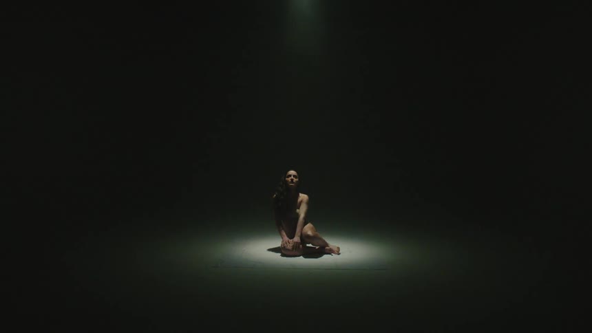 A woman lying on the floor in a dark room stands up as a spotlight shines on her and grows larger