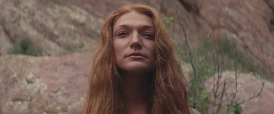 A woman with long red hair is standing outside with mountains behind her