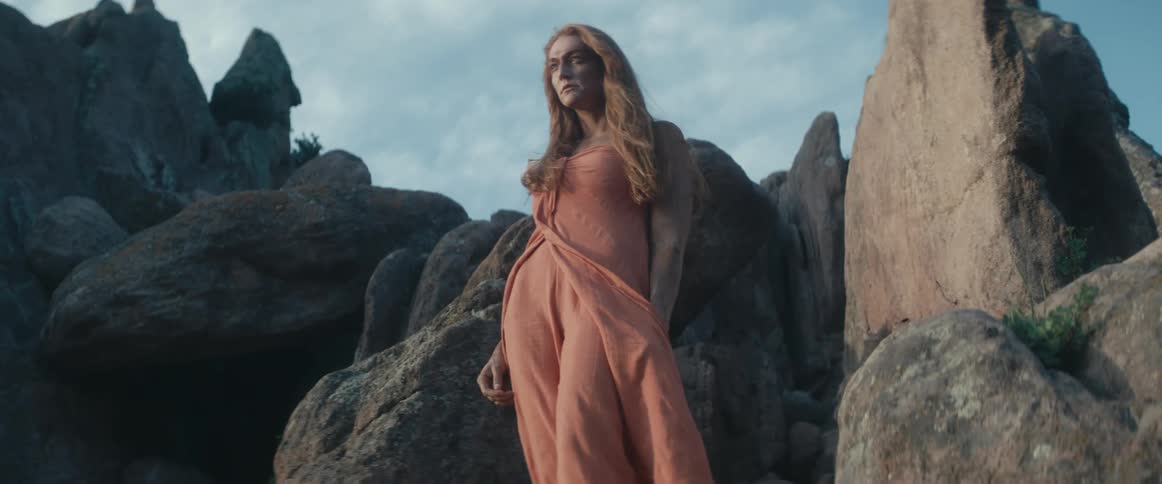 A barefoot woman with her skin covered in dirt stands atop a rock formation in the wilderness during the day while wearing a gown