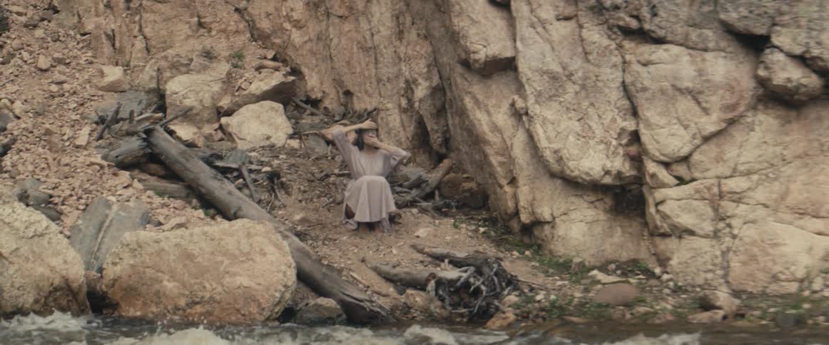 Woman crouches on river bank near rock wall covering her forehead and her mouth with her hands