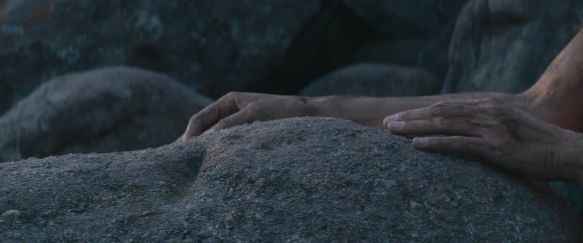 A woman covered in dirt looks around as she rubs a rock on a cliff