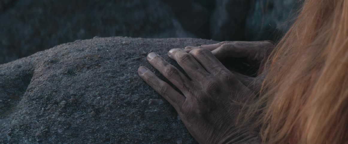 A woman covered in dirt stands by a canyon and runs her hands over a large rock
