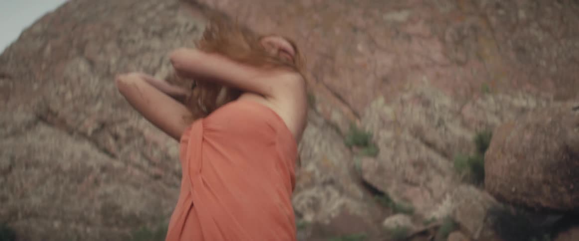 A woman standing at the base of a cliff picks up a large rock and throws it