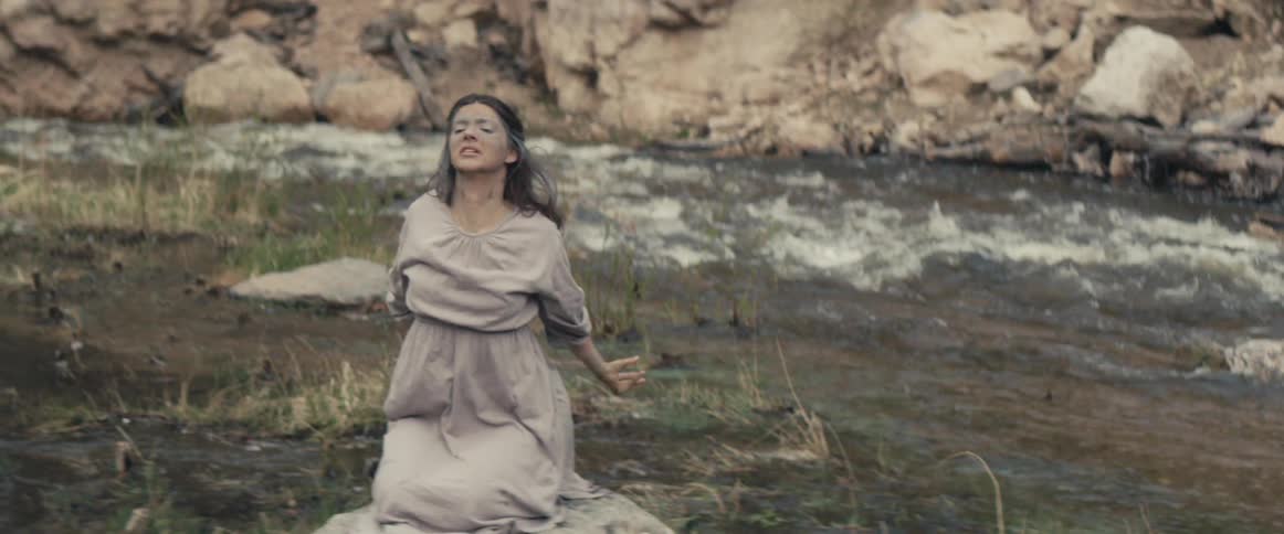 Woman with beige dress sits on rock and stretches her shoulders by the stream