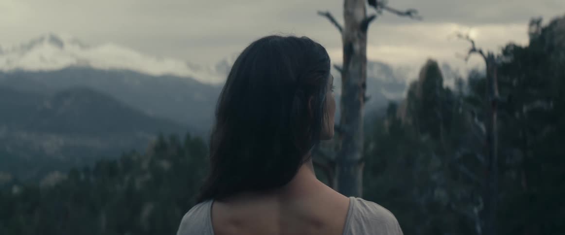 A woman stands in a forest area and looks towards a mountain range