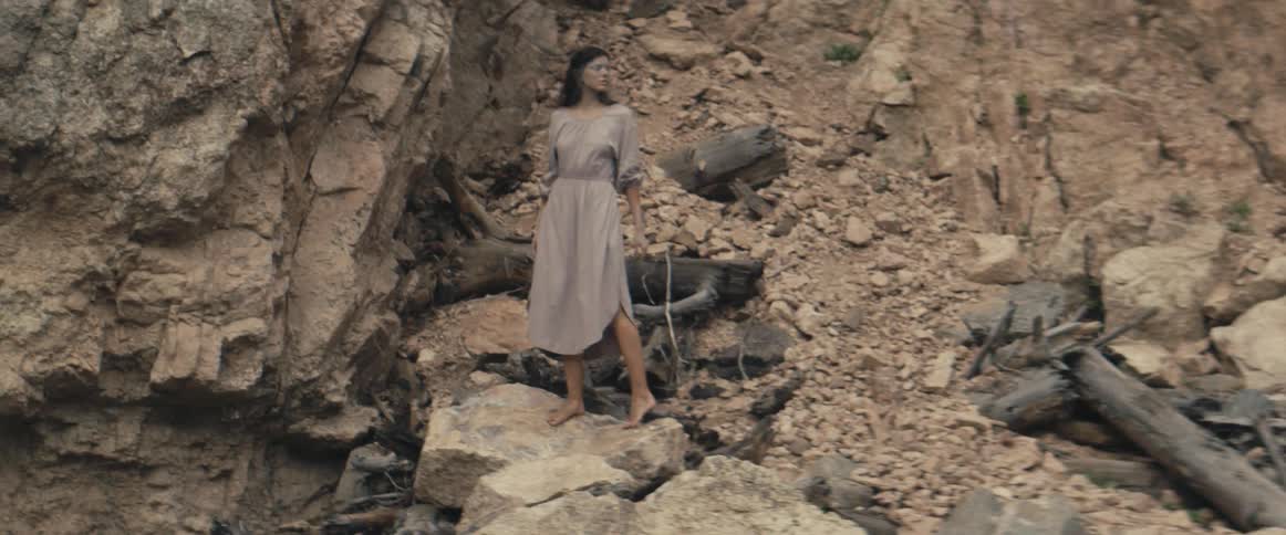 A woman wearing a gray dress walks barefoot up a mountain ledge