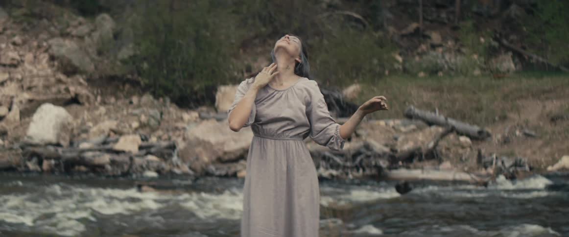 Woman with gray dress wavy hair and muddy face dances next to stream