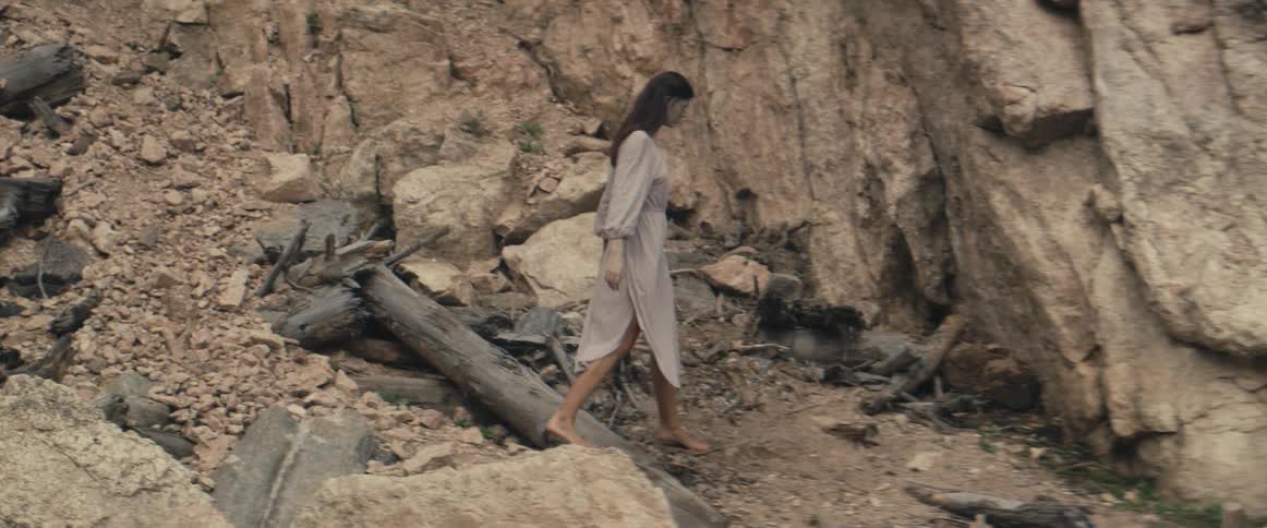 Woman with dark wavy hair and gray dress descends barefoot rocky hill
