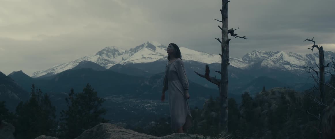 A woman wearing a gray dress walks barefoot up a ledge and looks at snow covered mountains in the distance