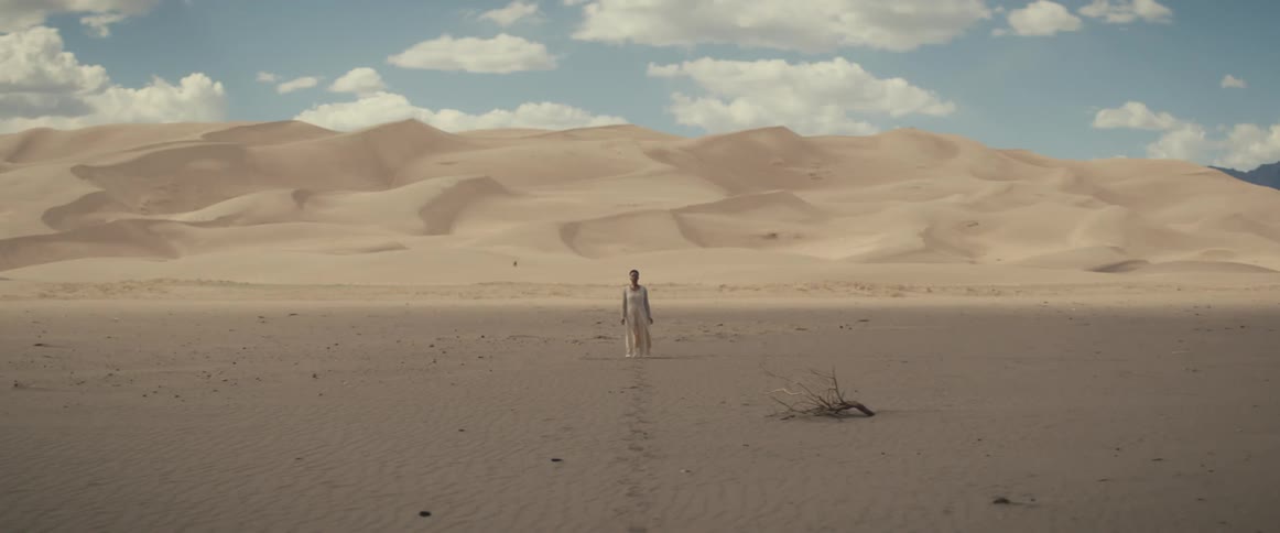 A woman in a long dress walks through the sandy desert under blue skies