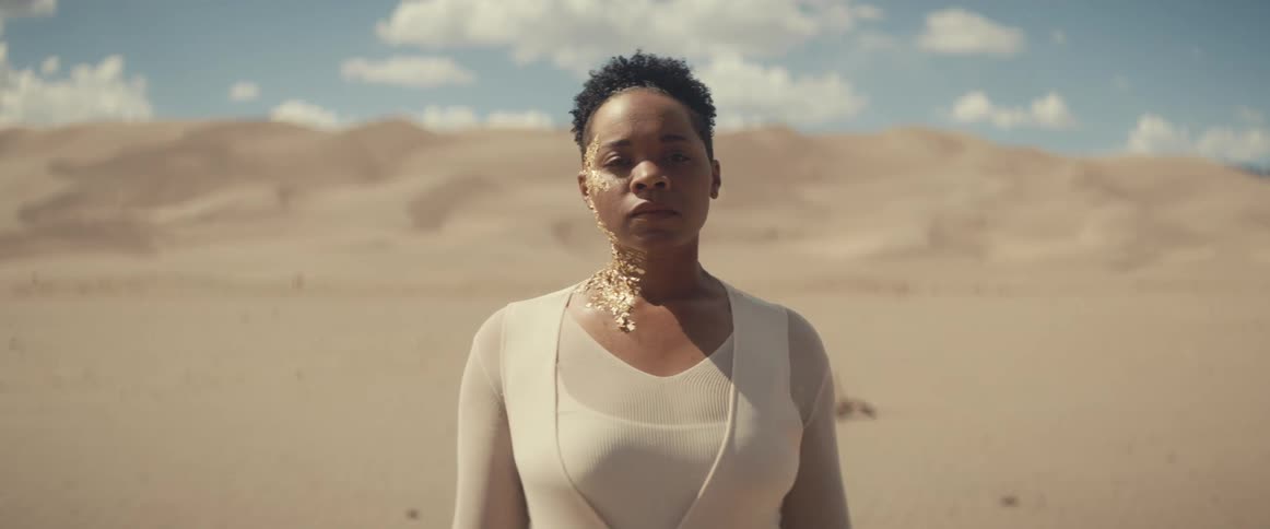 A woman walking through a desert with gold paint on one side of her head and neck comes to a stop before the camera