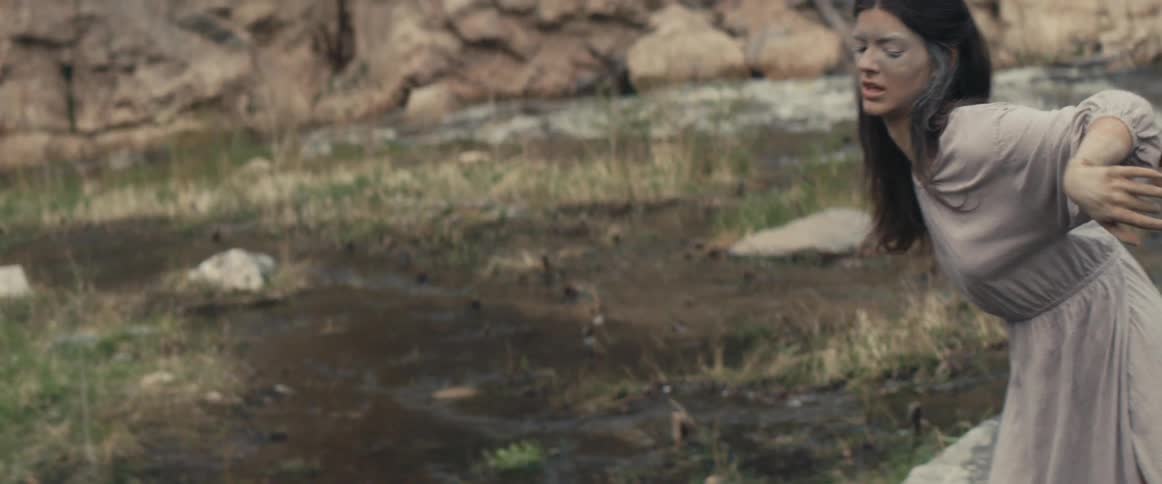 Woman with wavy hair and muddy face dances next to stream