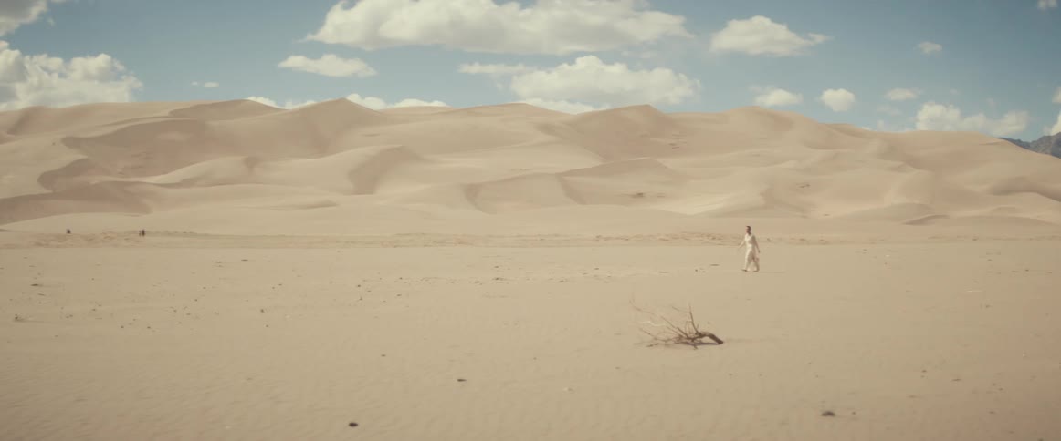 Someone wearing white clothes is walking across the desert under a cloud filled sky
