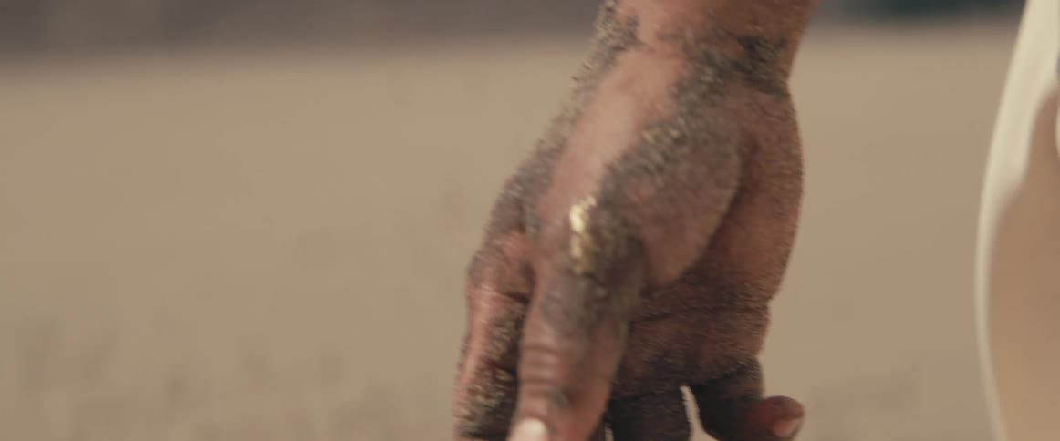 A woman walks in the desert with her arm covered in wet sand and some gold makeup on her thumb
