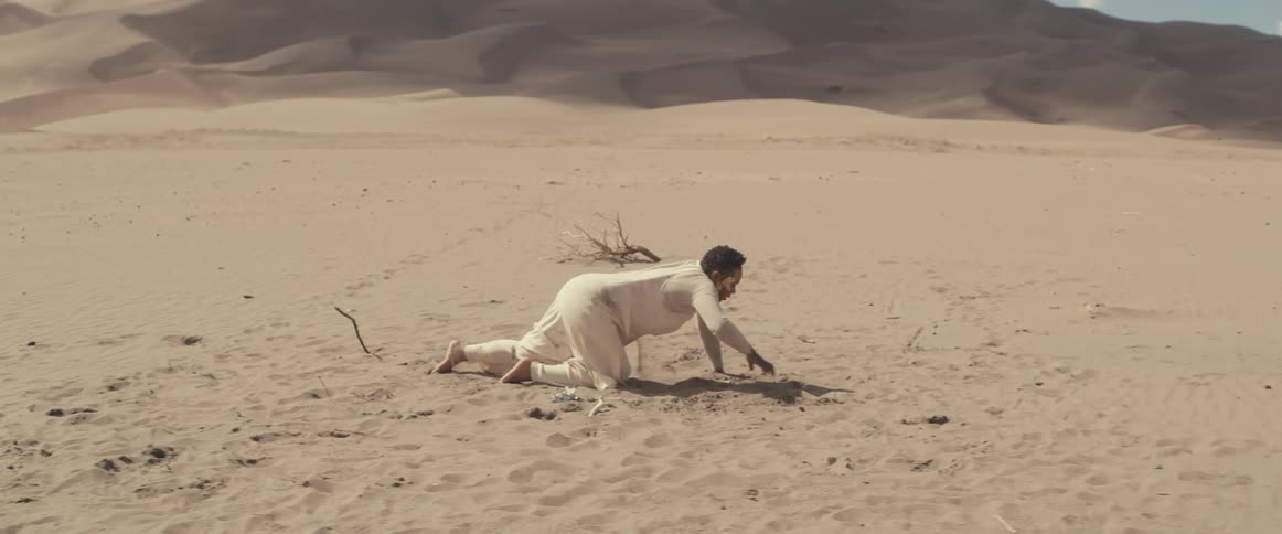 A woman wearing white is laying down on the desert sand and she slowly gets up and walks