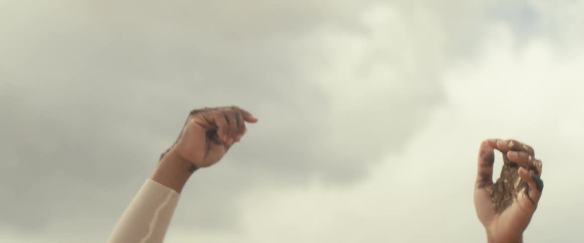 A black woman with tattoos on her hands and face reaches into the sky as she stands in the desert