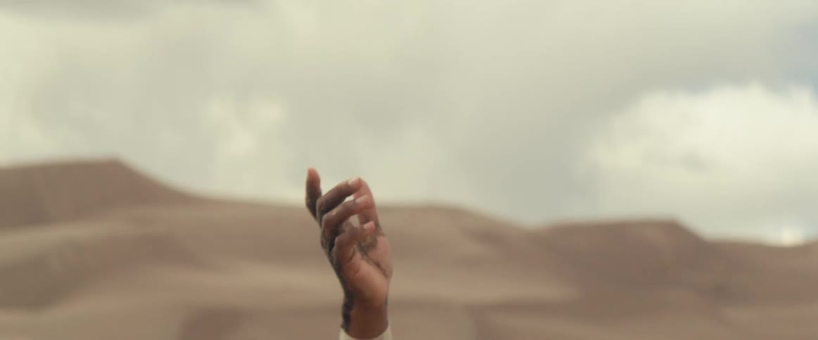 A woman with gold makeup on her face lifts her hands up towards the sky in the desert