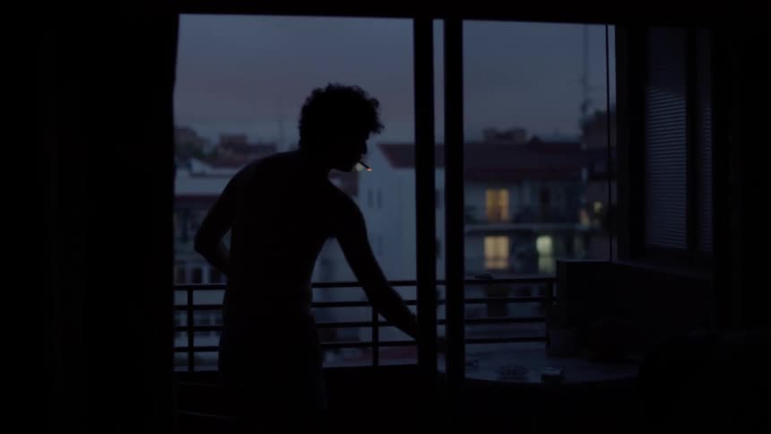 A man standing on his balcony lights a cigarette and leans on the railing and looks out across the street