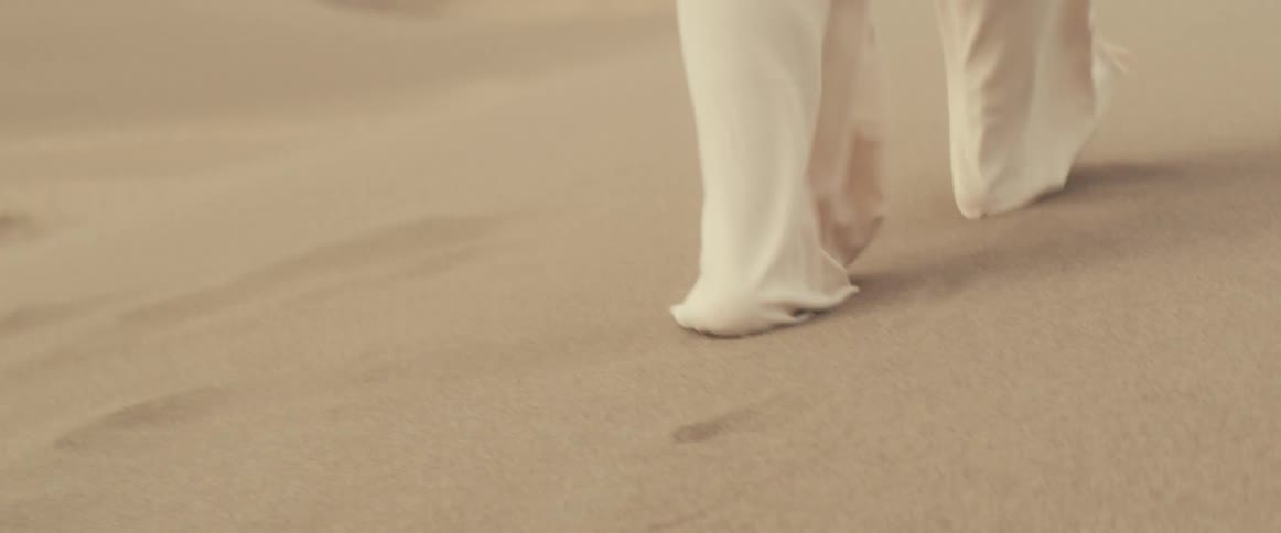 Someone wearing loose fitting white pants is walking barefoot through the desert