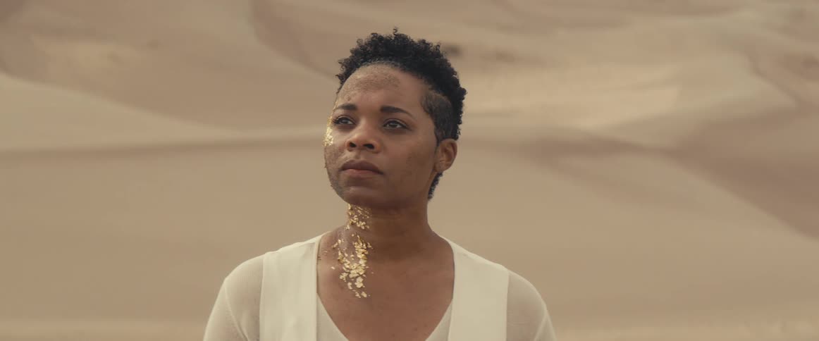A woman with gold makeup on one side of her face looks around in a contemplative manner