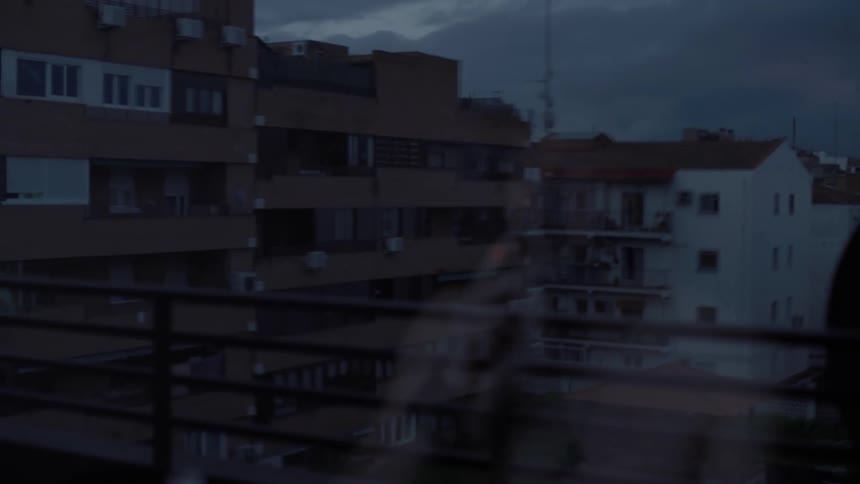 Shirtless mans lighting up cigarette is reflected on window glass watching apartment buildings in the city at dusk