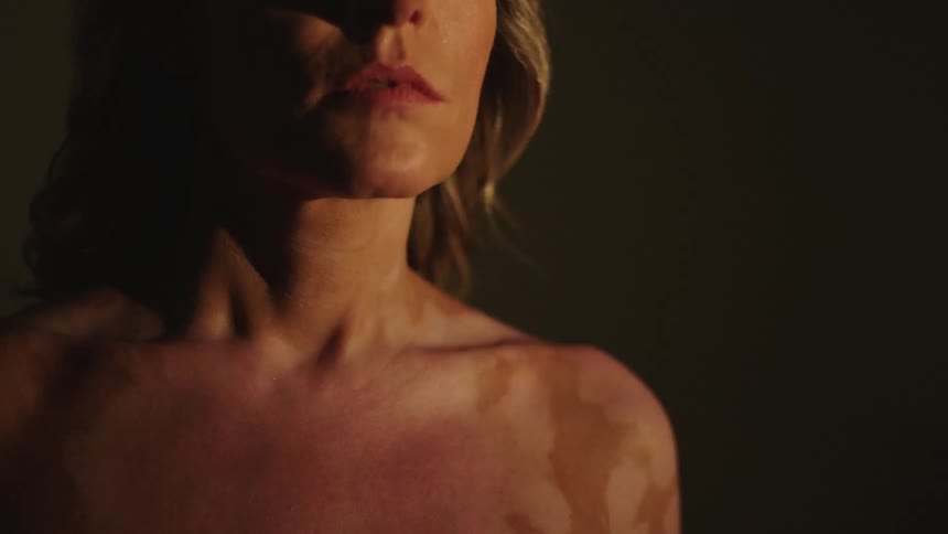 A woman stands in a white room as a light strobes over her bare upper chest
