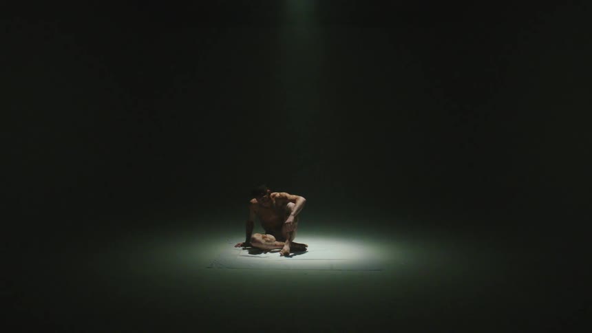 Spotlight turns on to reveal man with splotchy skin lying down on his side then he stands as light fills the room