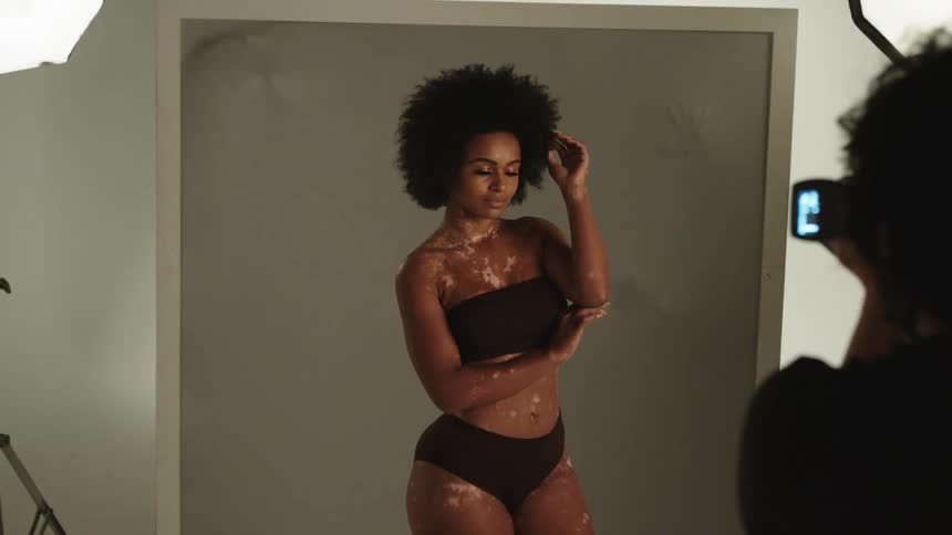 Model posing in a swimsuit during a photo shoot as hands push against the wall behind her