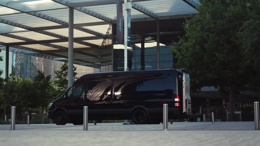 Male worker exits the driver's seat of a black transport van after parking in front of an office building in a city during the day