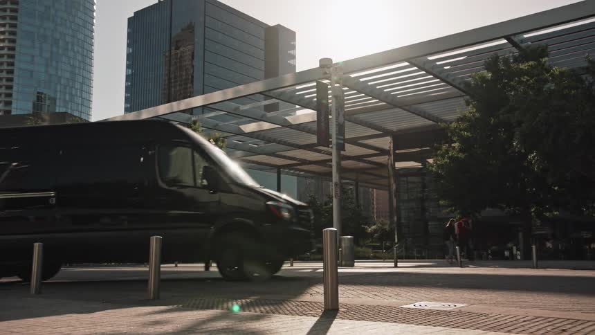 A large black transport van drives past the entrance to a building as people walk by