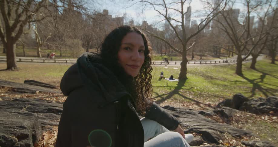 A woman sitting on a rock in a park turns and smiles softly
