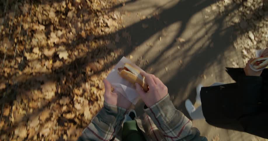 A man takes a bite of his hotdog as he walks on a park path with a friend