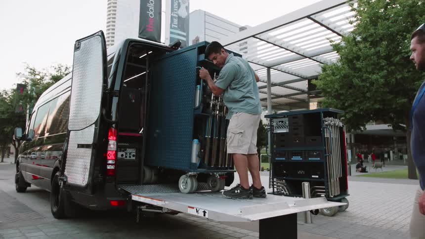 A man unloads equipment from the back of a van while another man watches from behind